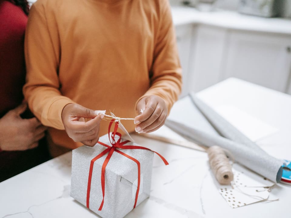 Ein Kind bindet mit Hilfe eines Erwachsenen eine rote Schleife um ein Geschenk